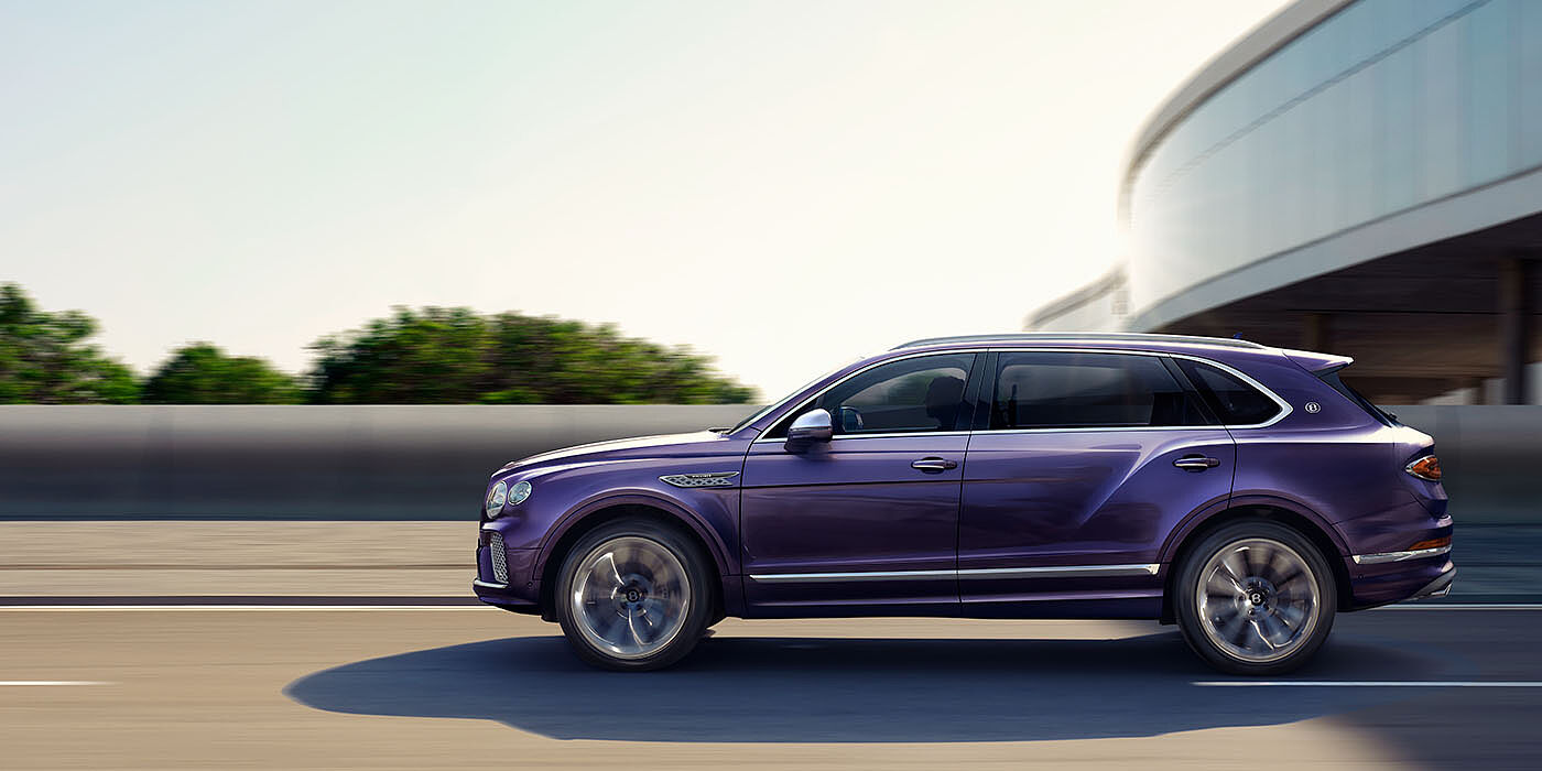 Bentley Glasgow Bentley Bentayga Extended Wheelbase Mulliner luxury SUV profile in Tanzanite Purple paint, driving on an country road past trees.