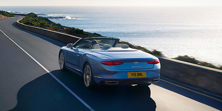 Bentley Glasgow New Bentley Continental GTC Azure convertible rear view in Light Windsor blue paint with 22 inch ten spoke wheels driving dynamically on an ocean road in the sunshine