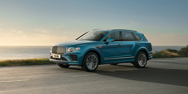 Bentley Glasgow Bentley Bentayga Azure SUV in Topaz blue paint driving dynamically by the ocean with 22 inch 10 spoke directional wheels