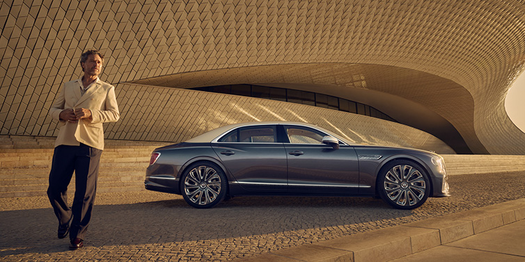 Bentley Glasgow Bentley Flying Spur Mulliner sedan in Rose Gold over Magnetic duotone paint parked by a curved wall with person in suit jacket standing on the left